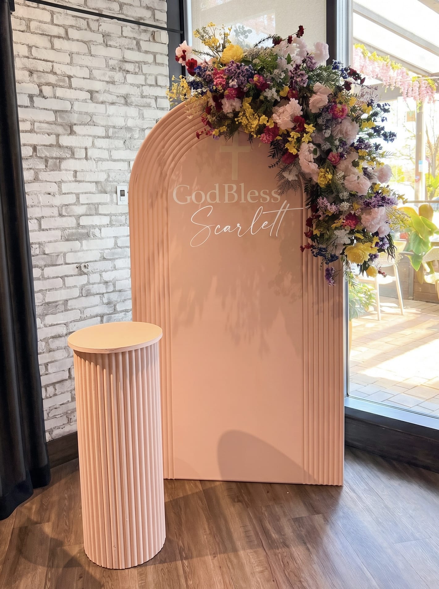 Blush ripple arch backdrop decorated with a colorful wildflower arrangement and custom blessing sign for a communion or baptism celebration.