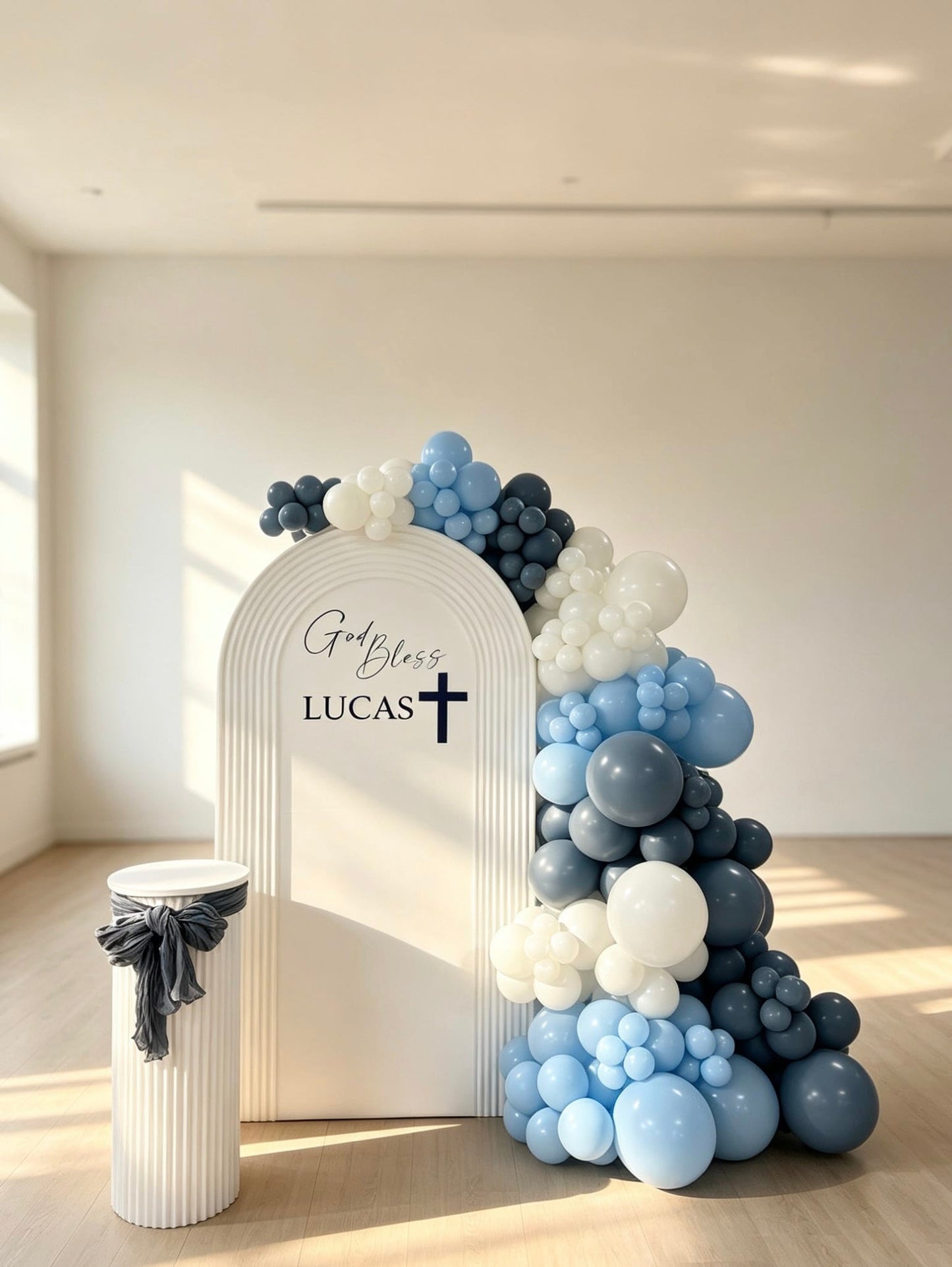 White ripple baptism arch backdrop with blue and white balloon garland, custom name, cross detail and matching white pedestal stand.