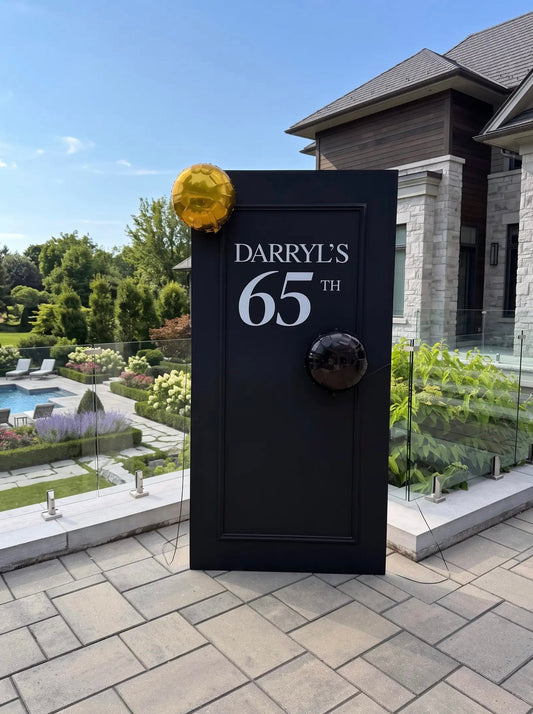 Black milestone birthday backdrop with framed panel detailing, custom white name and age lettering, and minimal gold and black balloon accents.