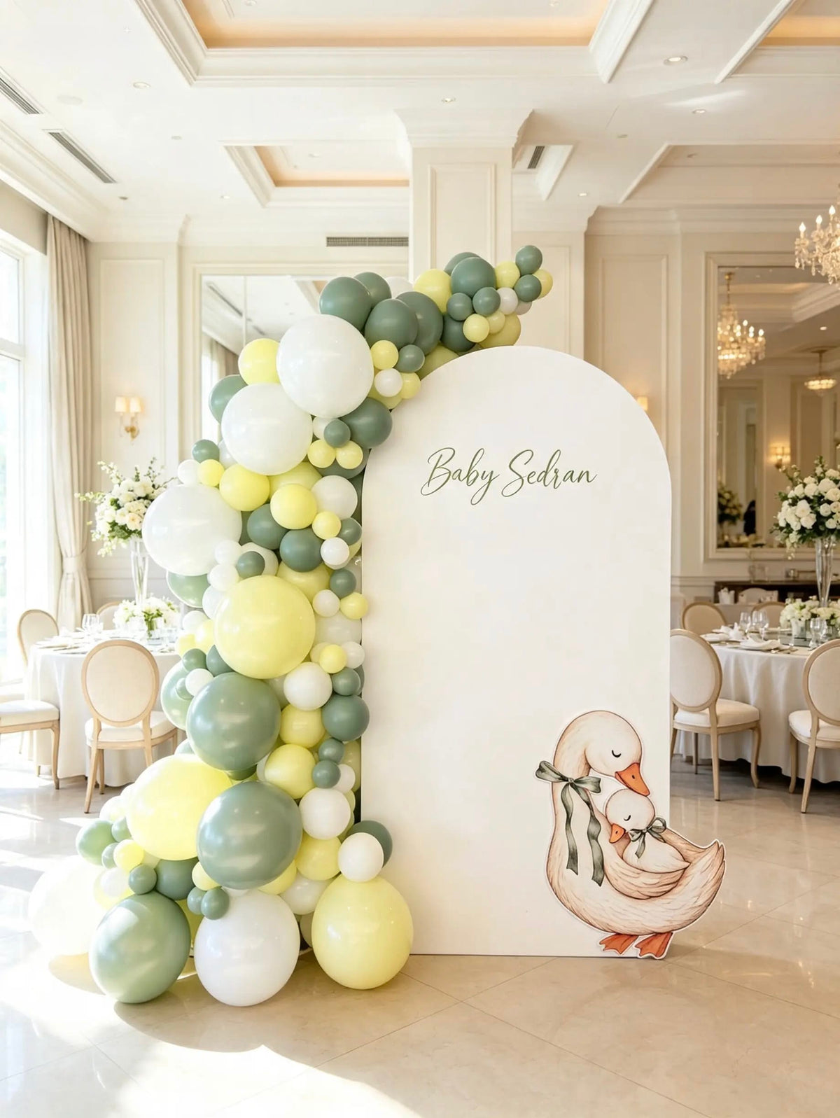 White baby shower arch with personalized lettering, sage green, yellow, and white balloons, and a mother-and-baby duck cutout.

