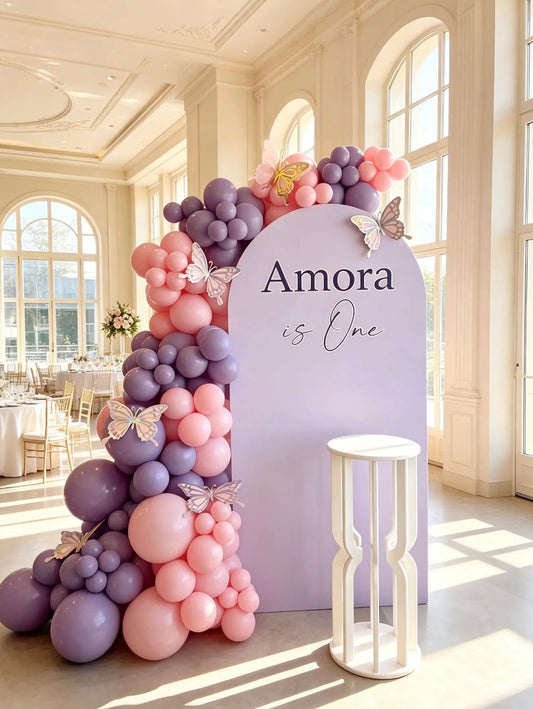 Lavender first birthday backdrop with pink and purple balloon garland, decorative butterflies, custom name lettering and white display stand.