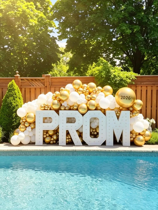 Outdoor prom display featuring illuminated PROM marquee letters with a white and metallic gold organic balloon installation and a personalized jumbo balloon.