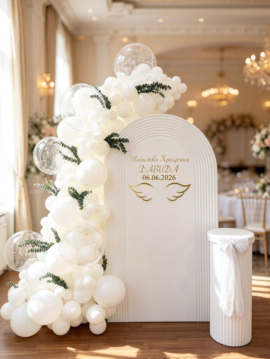 White baptism backdrop with a ripple arch, organic white balloon garland, clear bubble balloons, eucalyptus greenery, gold lettering and a matching pedestal stand.