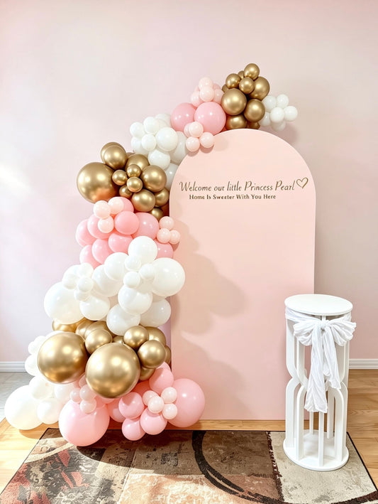 Blush pink welcome baby girl backdrop with white and gold balloon garland, custom arch backdrop and white pedestal stand with fabric bow.