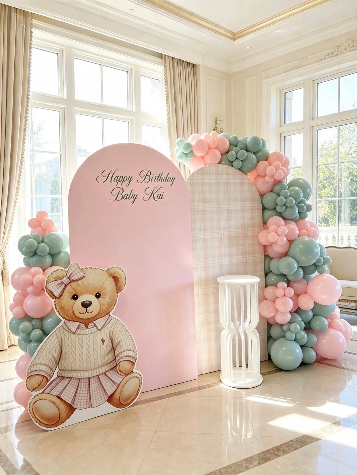 Pink and plaid birthday arches with personalized lettering, pink and sage balloons, a teddy bear cutout, and a white cake pedestal.