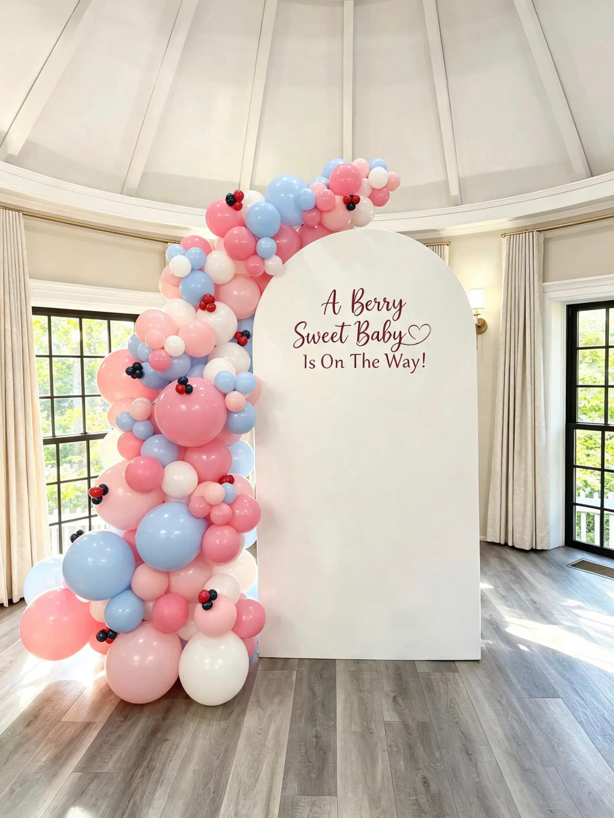 White arch with “A Berry Sweet Baby Is On The Way!” lettering, a pink, blue, and white balloon garland, and mini berry accents.