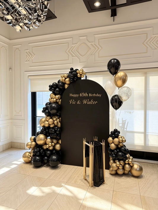 Elegant black and gold 65th birthday setup with black full arch backdrop, organic balloon garlands, helium balloons, and gold pedestal stand.