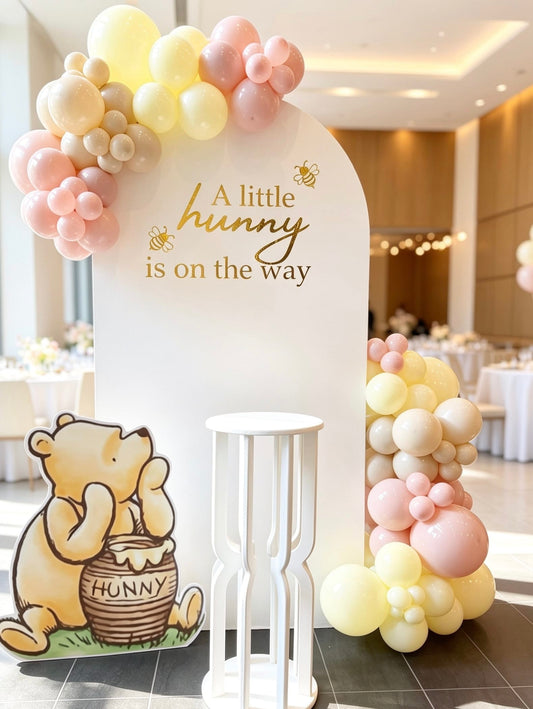 Classic Pooh baby shower backdrop with a white arch, blush pink, butter yellow and ivory balloon garlands, Pooh cutout and a white pedestal stand.