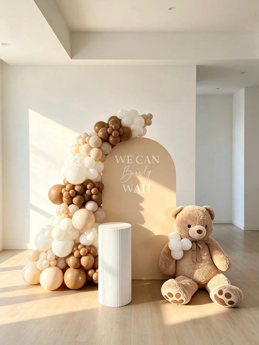 Neutral teddy bear baby shower backdrop with beige arch, cream and brown balloon garland, oversized teddy bear and white cake table.