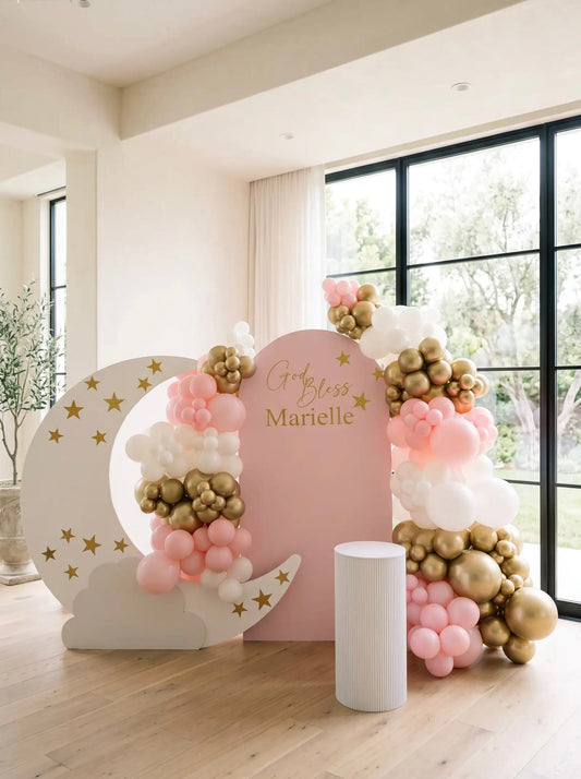 Pink baptism backdrop with a white crescent moon, gold stars, blush pink, white and gold balloon garland and personalized God Bless lettering.