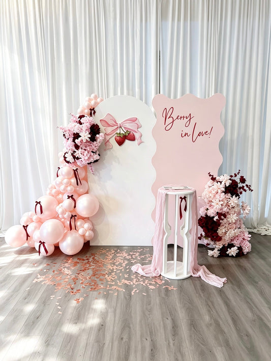Decorative setup with balloons, flowers, and a sign on a wooden floor.