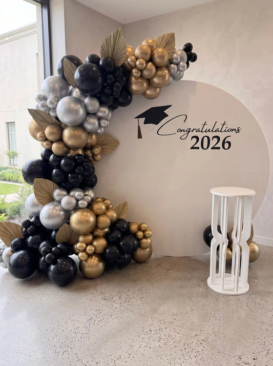 Black, gold and silver graduation backdrop with a large round panel, organic balloon garland, gold palm leaf accents and custom Congratulations lettering.