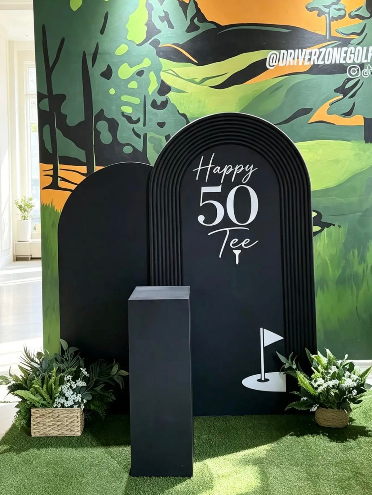 Black golf birthday arches with “Happy 50 Tee” lettering, golf graphics, a black cake pedestal, and greenery in woven baskets.