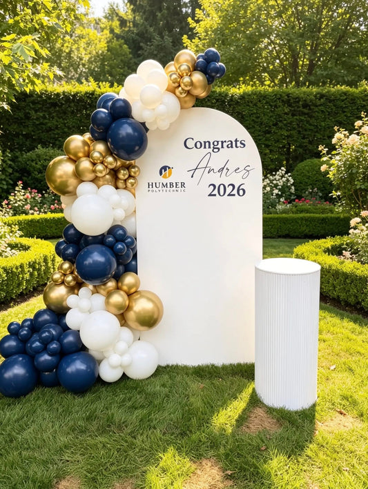 White graduation backdrop with navy blue, white and metallic gold balloon garland, personalized graduate name, school logo and white display stand.