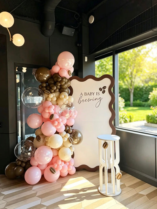 A Baby Is Brewing coffee theme baby shower backdrop with pink, cream and brown balloon garland, clear bubble balloons, coffee bean accents and white cake table.
