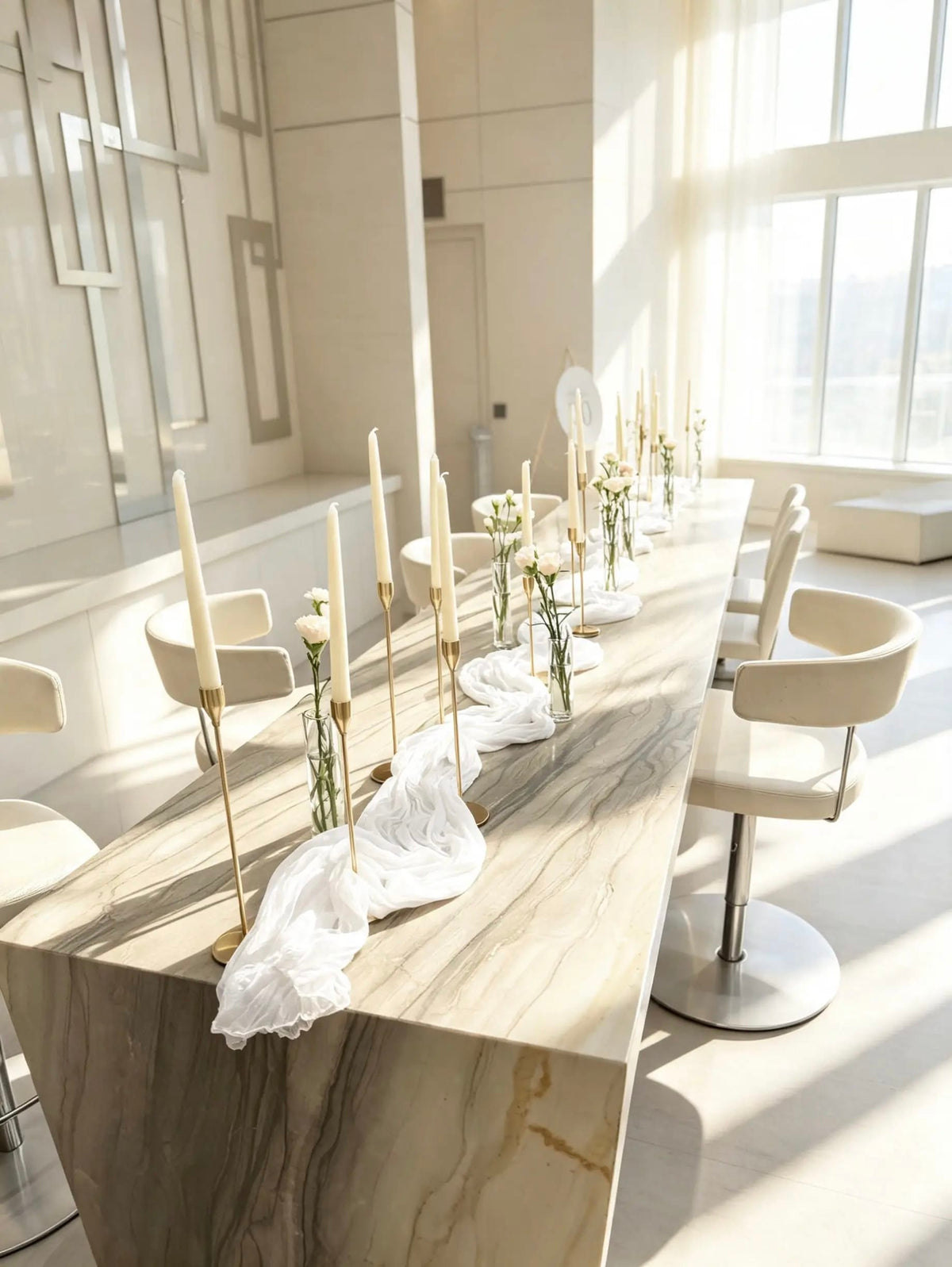 Elegant table decor with ivory taper candles in gold holders, white flowers in clear bud vases, and a draped white runner.