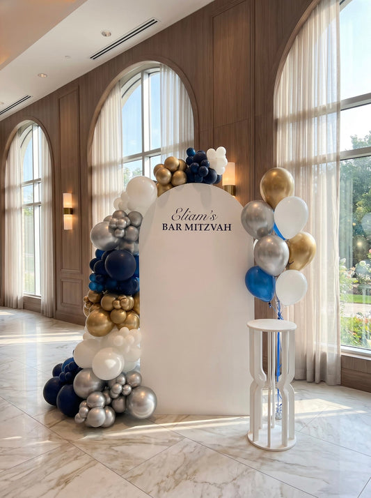 White Bar Mitzvah backdrop with navy, gold, silver and white balloon garland, custom name sign and matching helium balloon bouquet.