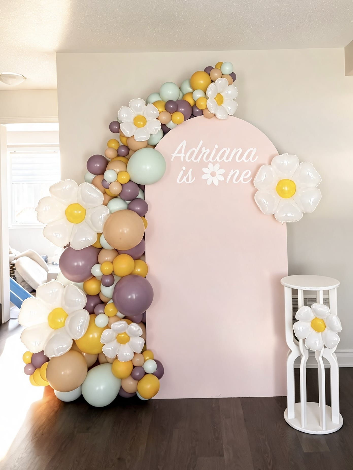 Pink daisy birthday backdrop with pastel balloon garland, oversized flower balloons and custom first birthday sign for a baby girl celebration.