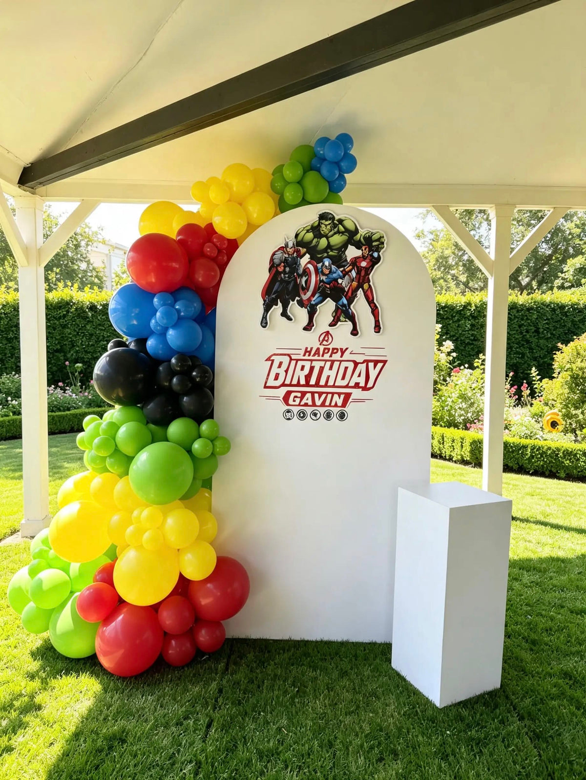White Avengers birthday arch with superhero characters, personalized lettering, a colourful balloon garland, and a white cake pedestal.