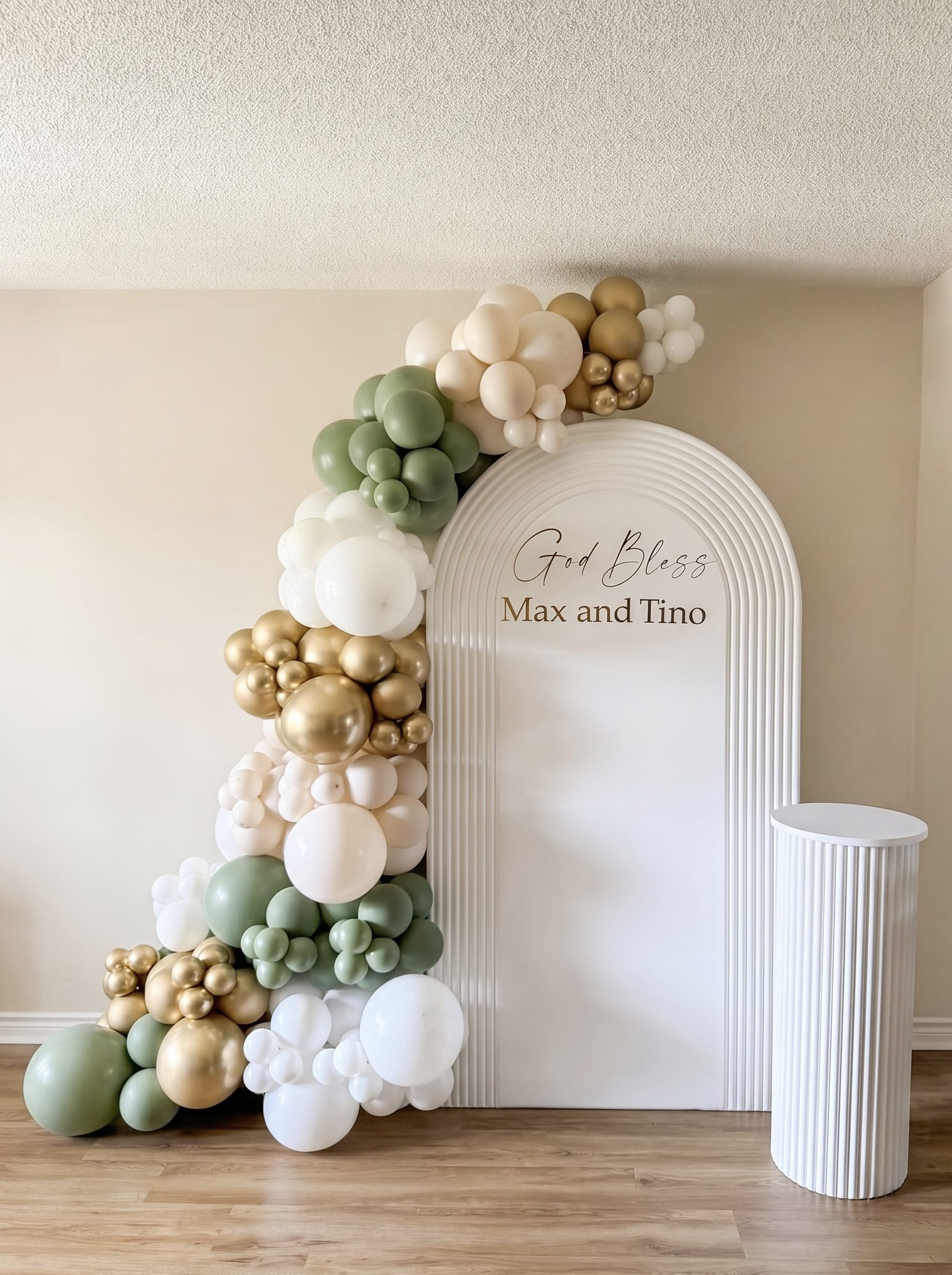 White ripple arch backdrop with sage green, white and gold balloon garland, custom gold lettering and matching pedestal stand.