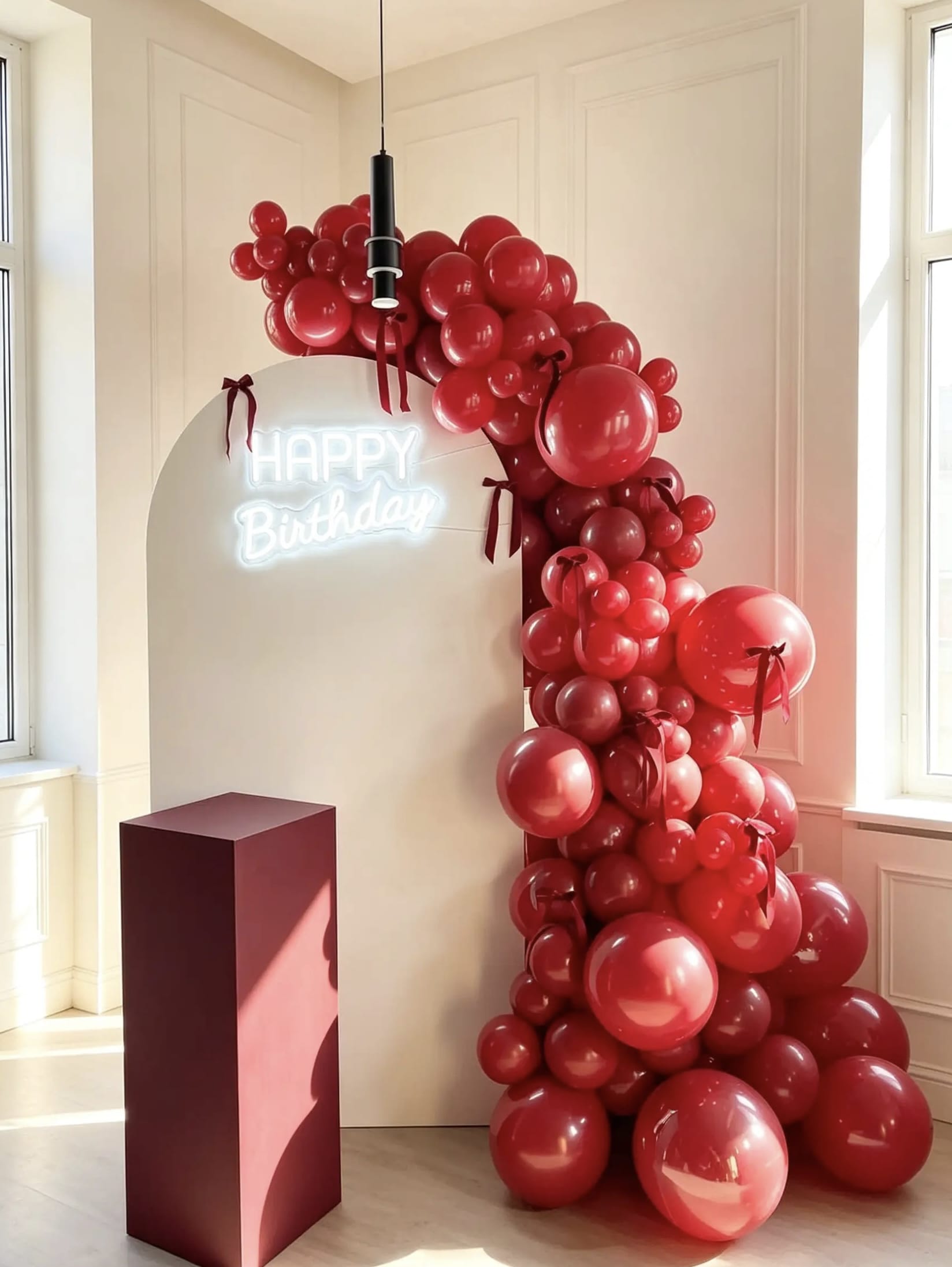 White arch birthday backdrop with red balloon garland, red bow accents, Happy Birthday neon sign and burgundy pedestal