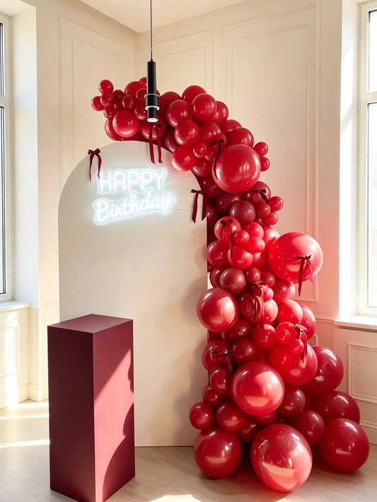 White arch birthday backdrop with red balloon garland, red bow accents, Happy Birthday neon sign and burgundy pedestal