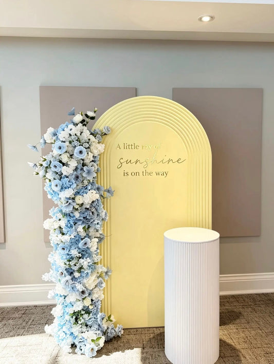 Yellow ripple arch baby shower backdrop with a blue and white floral installation and a white ribbed cake pedestal.