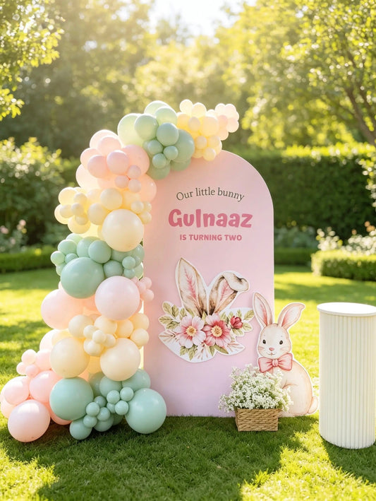Little bunny birthday backdrop with pink arch, pastel pink, sage green and ivory balloons, bunny cut-outs and white ribbed cake table.