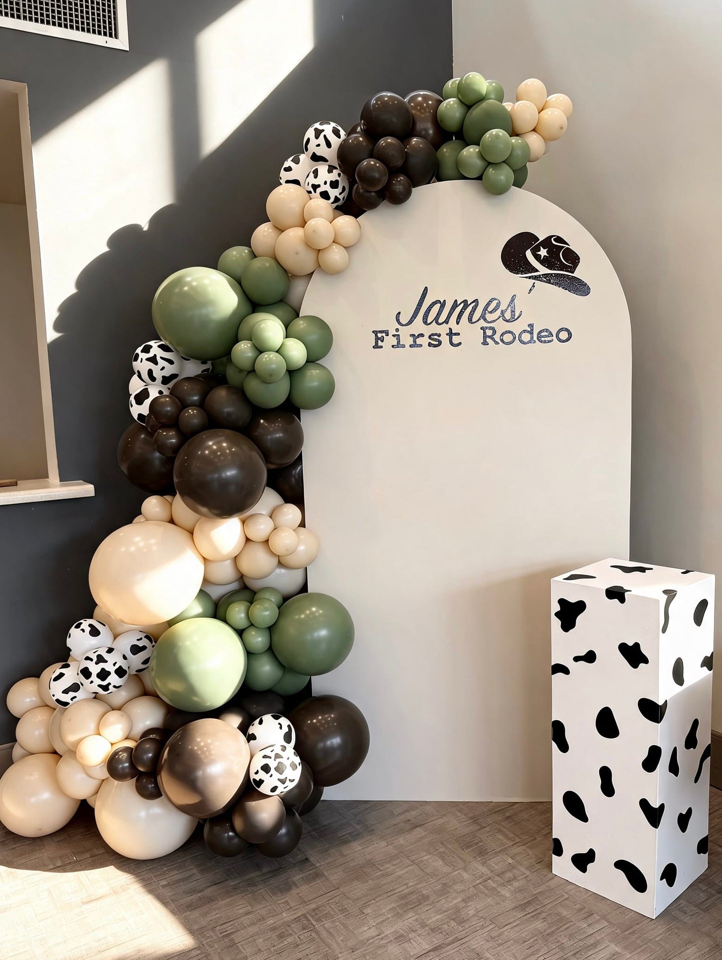 Western rodeo birthday backdrop with white arch panel, sage green and brown balloon garland, cow print accents and matching cow print pedestal stand.