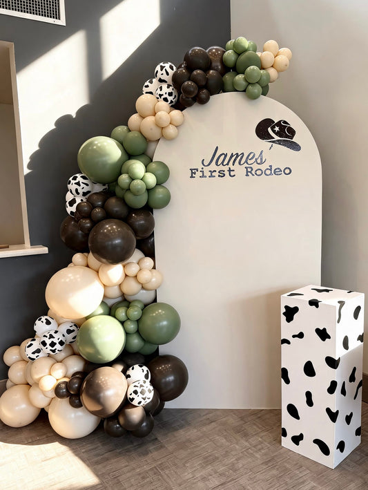 Western rodeo birthday backdrop with white arch panel, sage green and brown balloon garland, cow print accents and matching cow print pedestal stand.