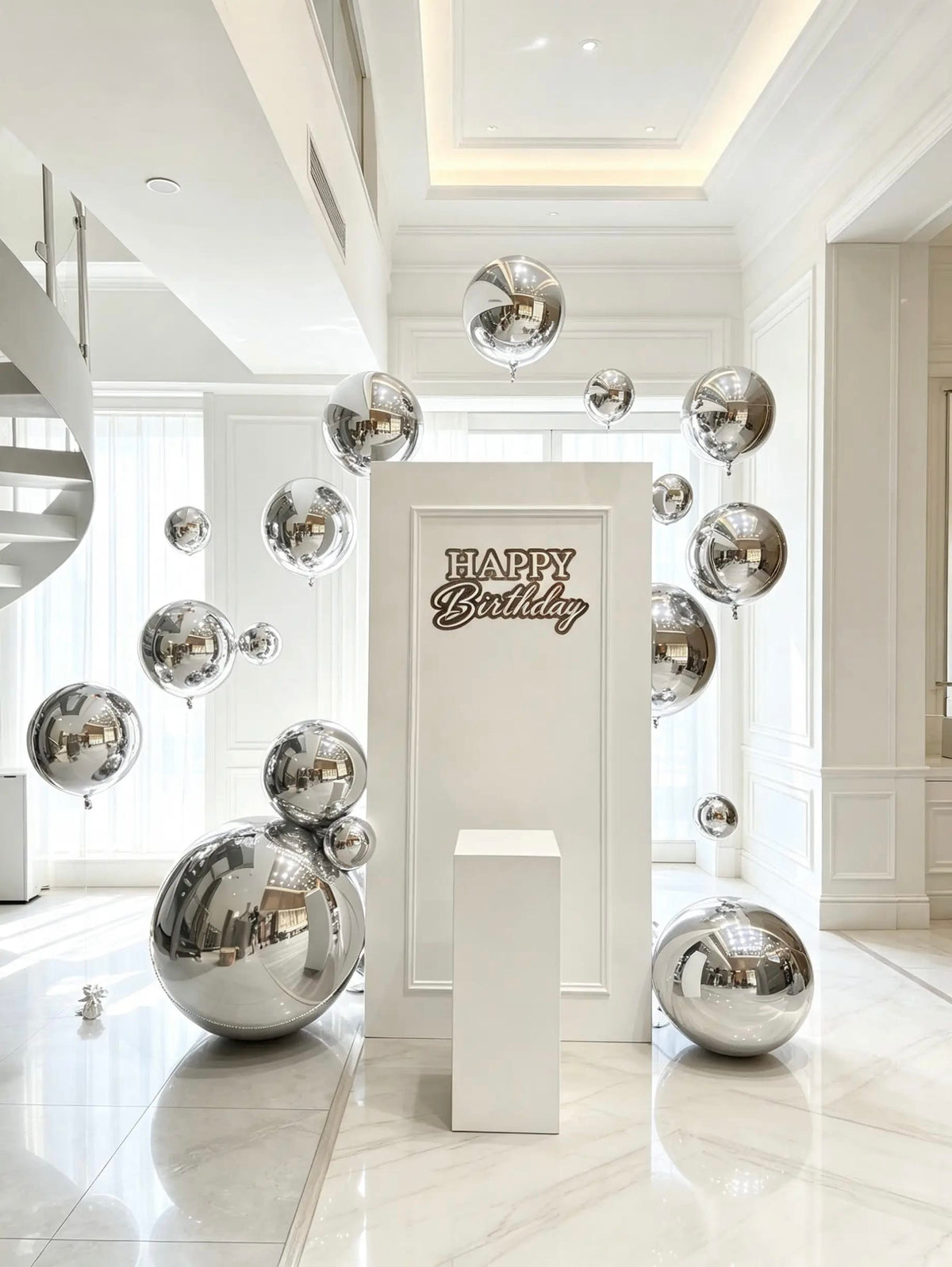White birthday backdrop with a “Happy Birthday” sign, reflective silver sphere balloons in assorted sizes, and a white cake pedestal.