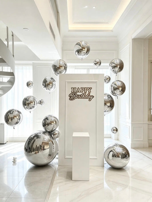 White birthday backdrop with a “Happy Birthday” sign, reflective silver sphere balloons in assorted sizes, and a white cake pedestal.