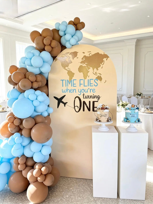 Travel-themed first birthday backdrop with a beige arch, baby blue and mocha balloon garland, world map design and two white pedestal stands.