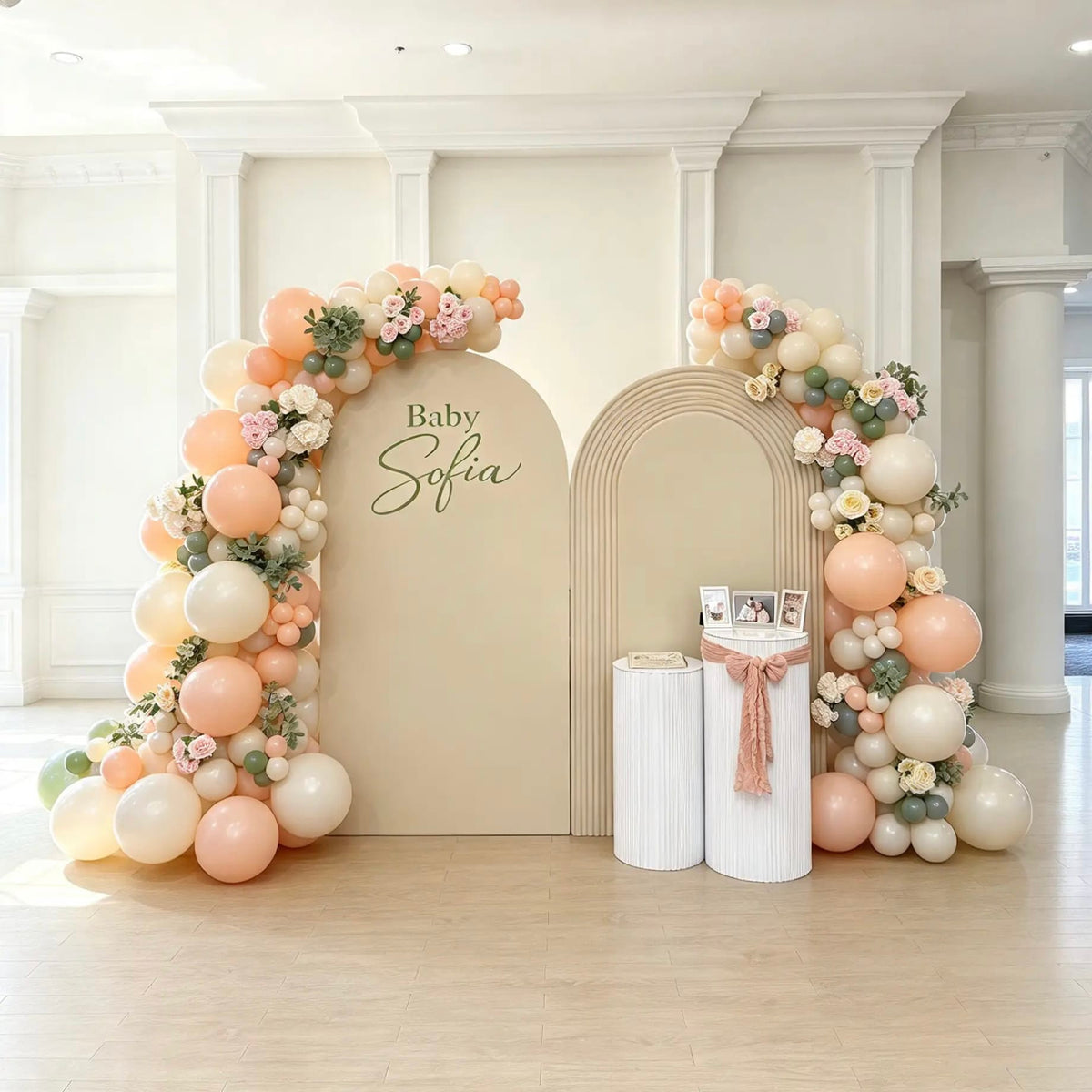 Baby shower backdrop with two beige and ivory arches, personalized Baby Sofia lettering, peach and sage green balloon garlands, pink and white floral accents, and two white ribbed pedestals.