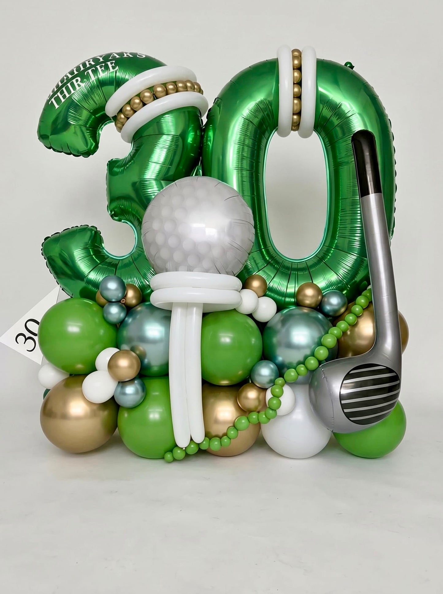 Golf-themed milestone birthday balloon stand featuring oversized green number balloons, a custom golf ball centerpiece, giant golf club accent and personalized details in green, gold and white.