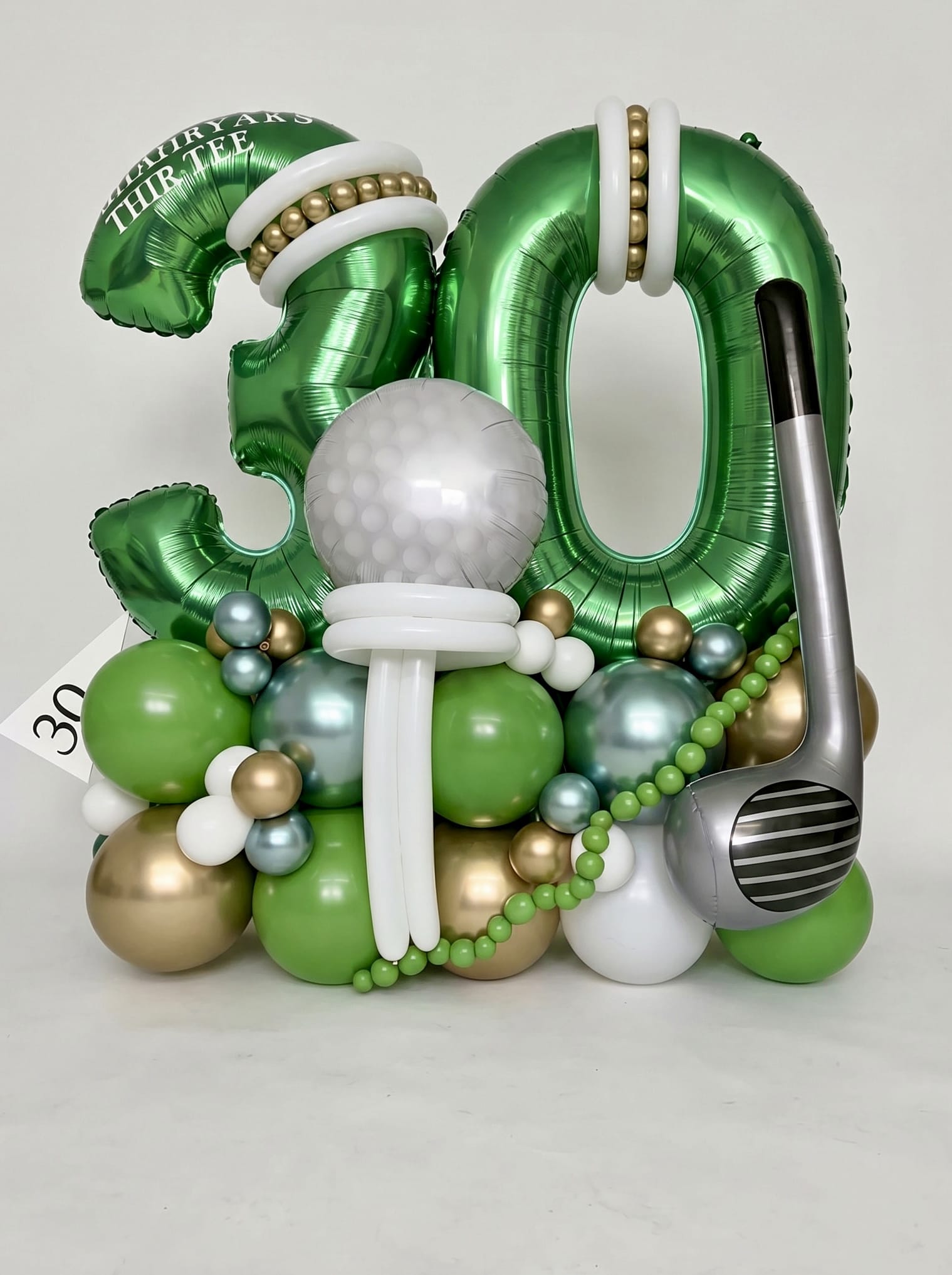 Golf-themed milestone birthday balloon stand featuring oversized green number balloons, a custom golf ball centerpiece, giant golf club accent and personalized details in green, gold and white.