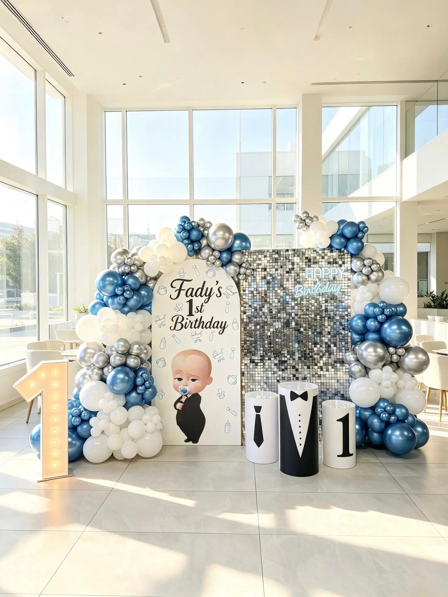 Boss Baby first birthday backdrop with blue, white and silver balloons, silver shimmer wall, themed cake tables and marquee number 1.
