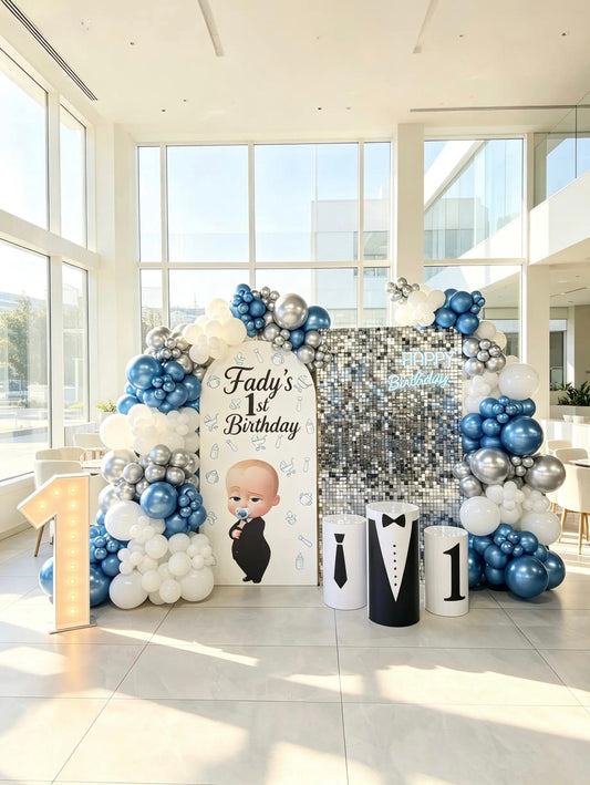 Boss Baby first birthday backdrop with blue, white and silver balloons, silver shimmer wall, themed cake tables and marquee number 1.