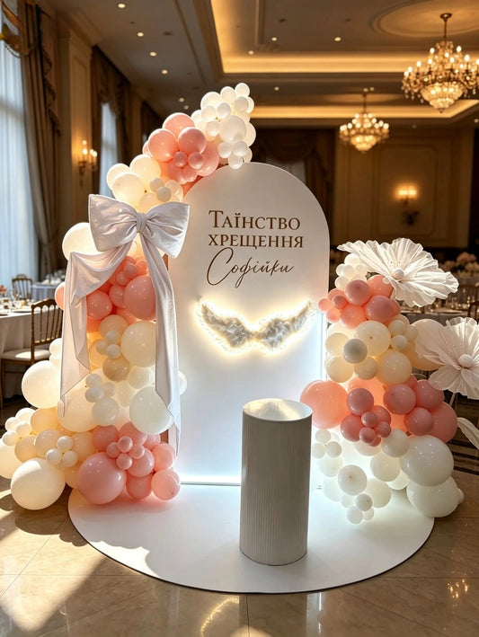 Pink and white christening balloon backdrop with personalized arch, giant flowers, oversized bow, illuminated angel wings and white cake table.