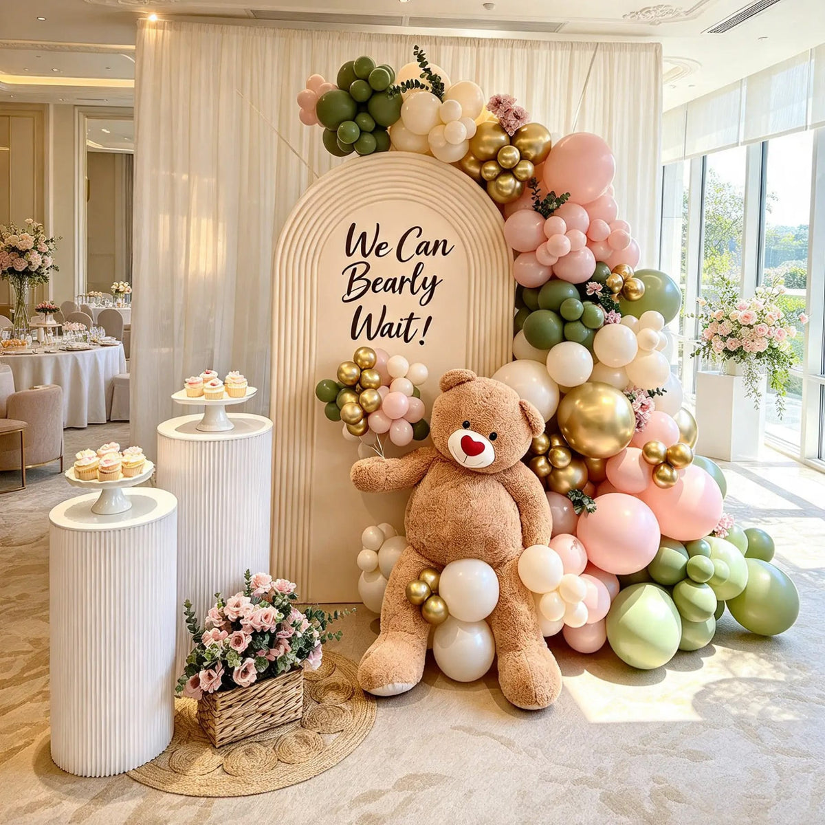 Cream “We Can Bearly Wait” arch with pink, green, and gold balloons, a teddy bear, white dessert pedestals, and a floral basket.