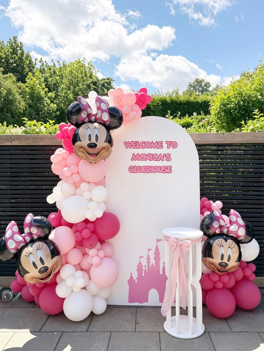 Minnie Mouse birthday backdrop with a white arch, pink balloon garland, three Minnie Mouse foil balloons, custom lettering and a white pedestal stand.
