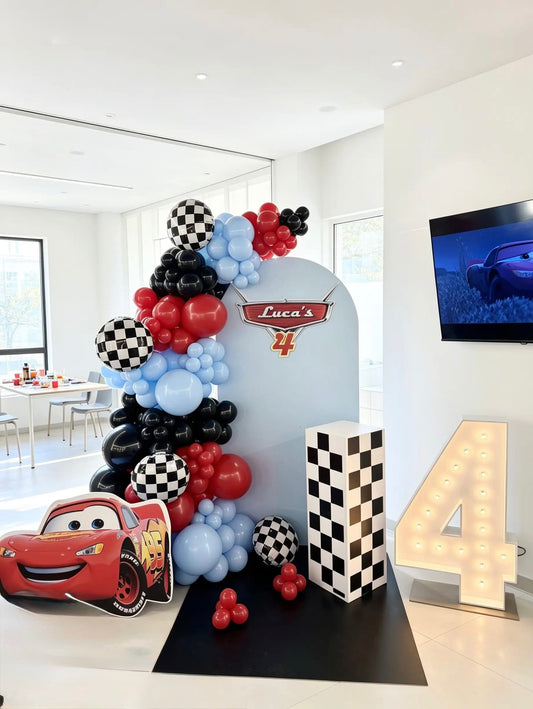 Cars-themed 4th birthday backdrop with red, black and blue balloons, checkered racing details, Lightning McQueen cutout and light-up marquee number 4.