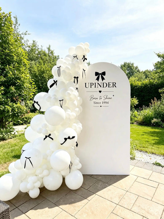 White birthday arch with personalized “Born to Shine” lettering beside an organic white balloon garland decorated with black ribbon bows.
