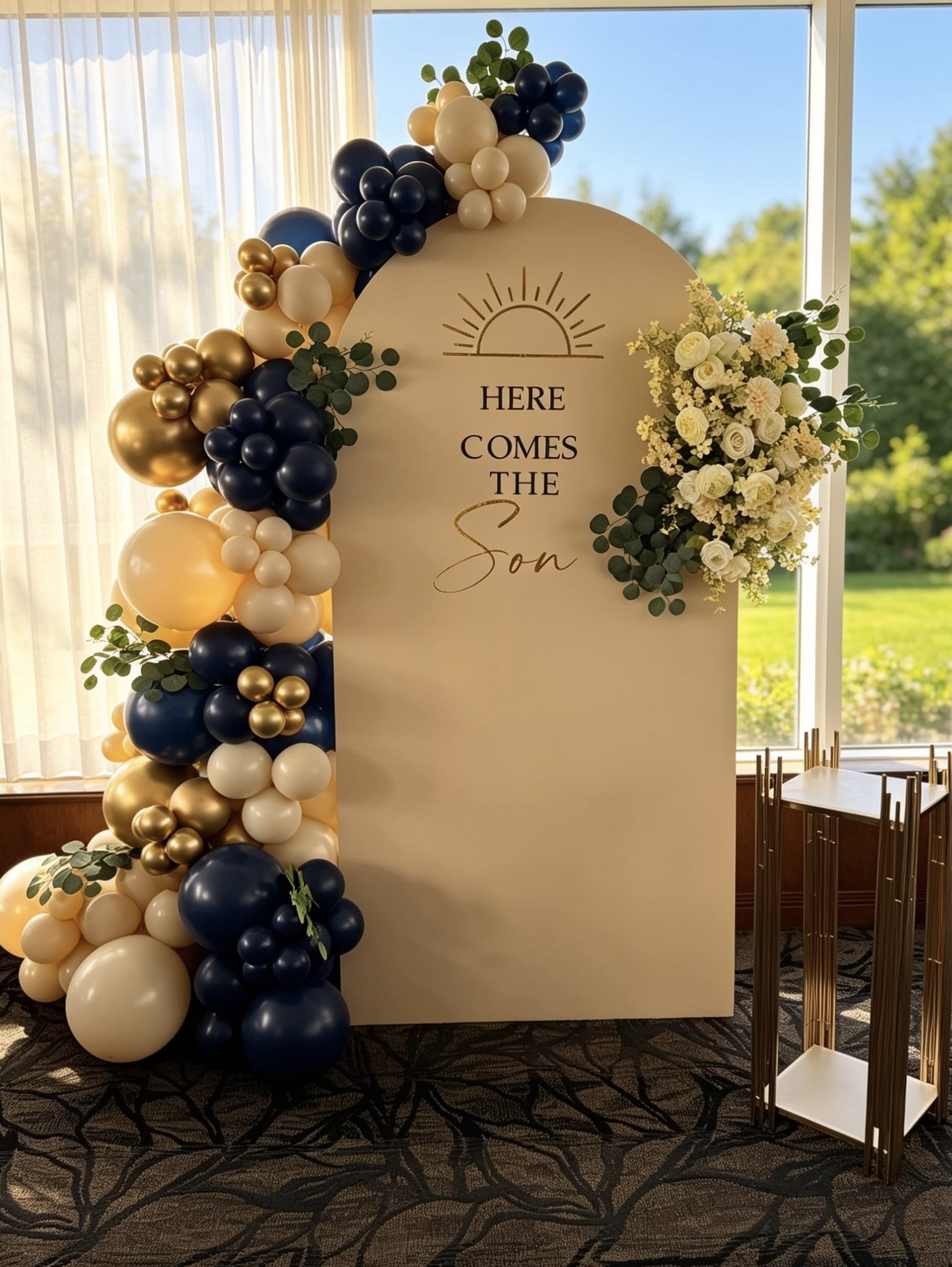 Beige baby shower backdrop with a navy blue, ivory and gold balloon garland, greenery, coordinating floral arrangement and a modern gold display stand.