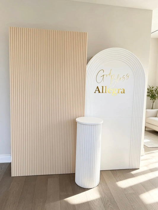 Beige ribbed backdrop and white ripple arch with personalized gold God Bless lettering and a white ribbed pedestal for a baptism celebration.