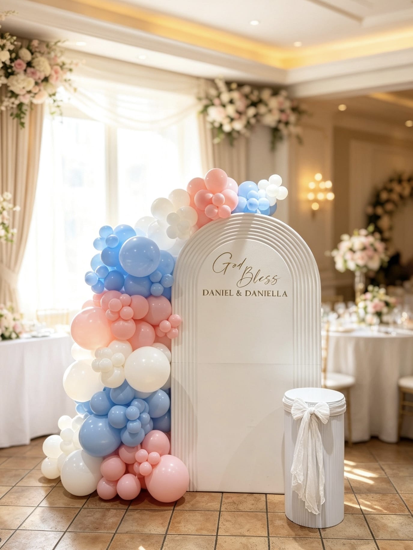 Twin baptism backdrop with a white ripple panel, blush pink, baby blue and white balloon garland, custom names and matching pedestal.