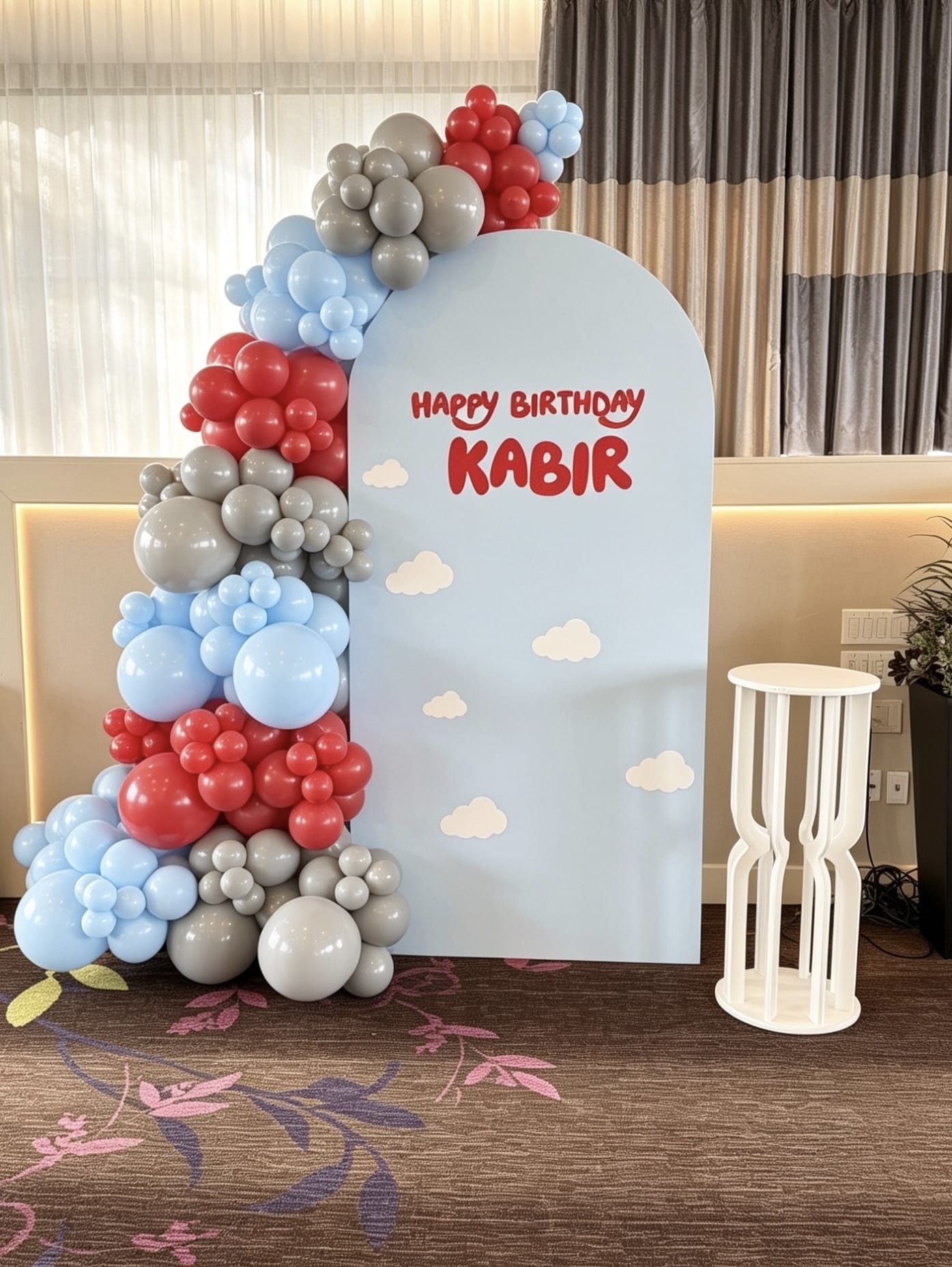 Light blue birthday backdrop with cloud accents and blue, red and grey balloon garland for a kids celebration.