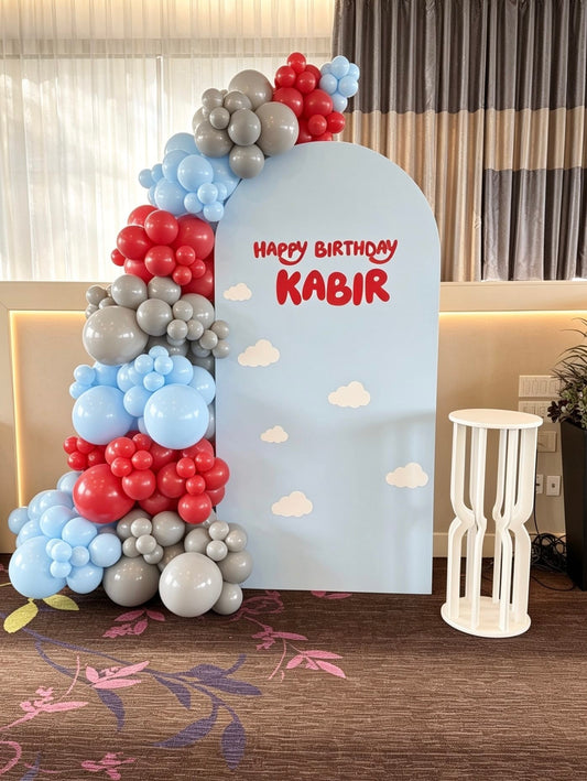 Light blue birthday backdrop with cloud accents and blue, red and grey balloon garland for a kids celebration.