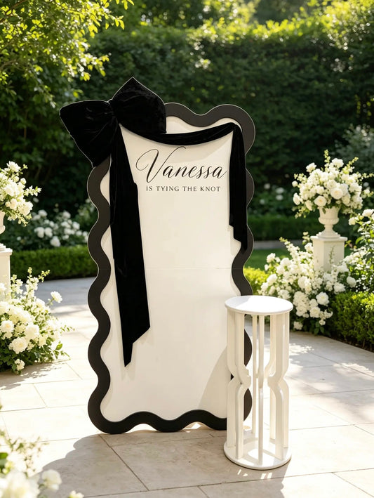 Black and white bridal shower wave backdrop with personalized name, oversized black velvet bow, velvet draping and white cake table.