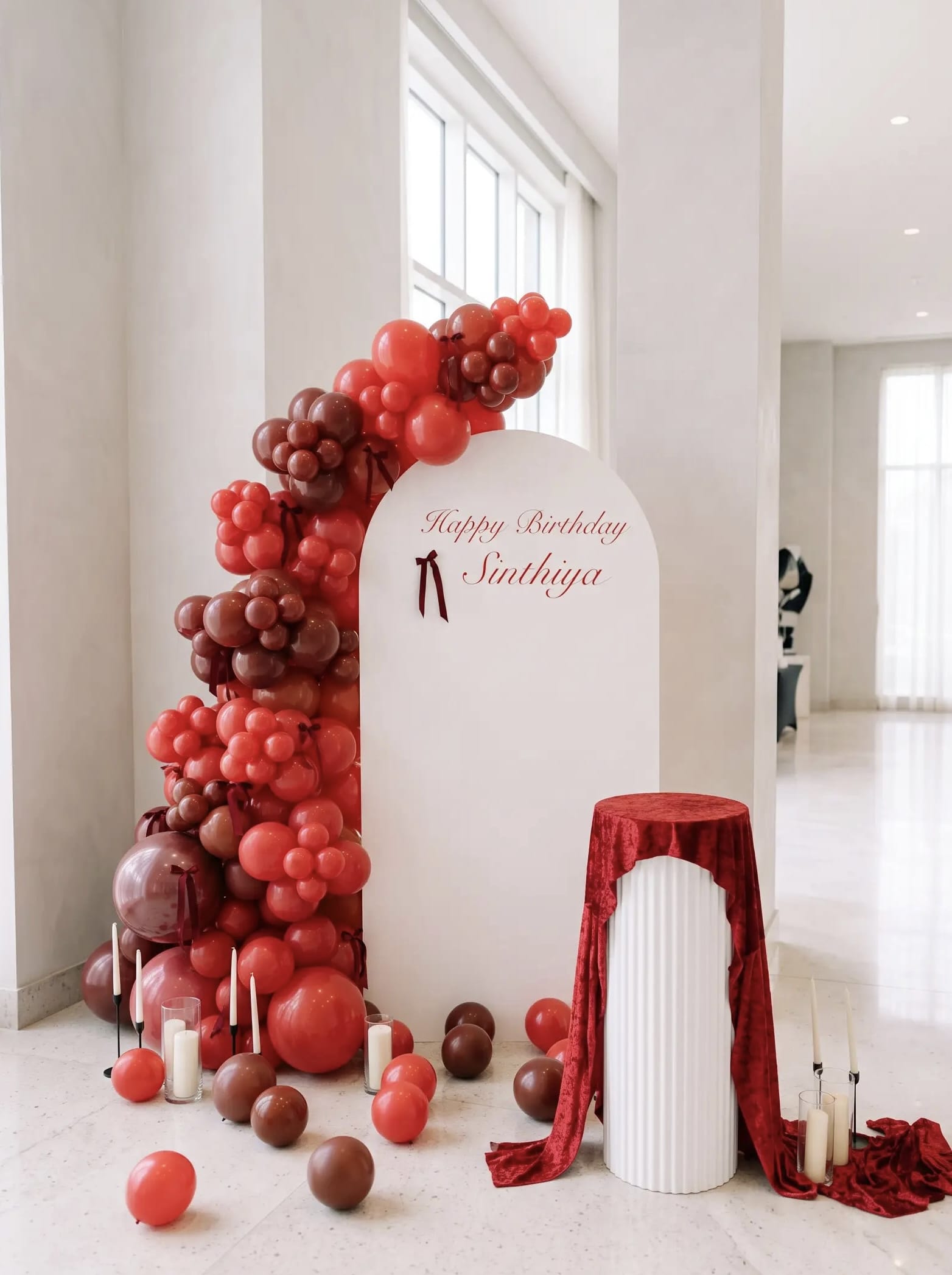 White birthday backdrop with red and burgundy balloon garland, bow accents, personalized lettering, candles and a red velvet-draped display stand.